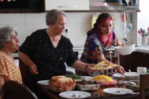 Three women enjoy serving a homemade meal in a lively kitchen setting.